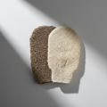 A pair of exfoliating gloves, one with a brown textured surface and another with a beige, sponge-like appearance, placed on a two-tone background with pink and white colors.