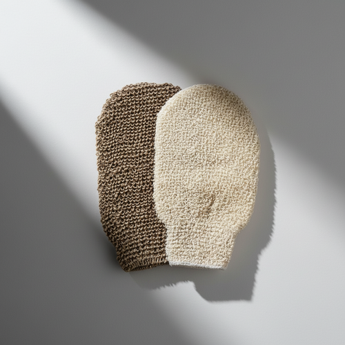 A pair of exfoliating gloves, one with a brown textured surface and another with a beige, sponge-like appearance, placed on a two-tone background with pink and white colors.